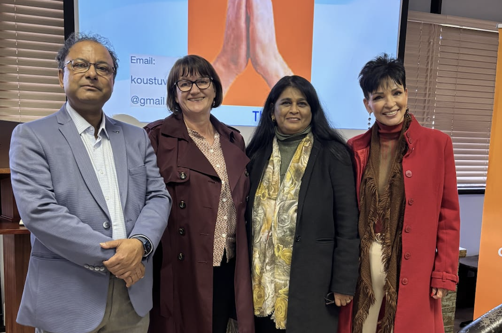 Four adults stand side by side indoors, smiling for a group photo in front of a screen with blue and orange graphics.